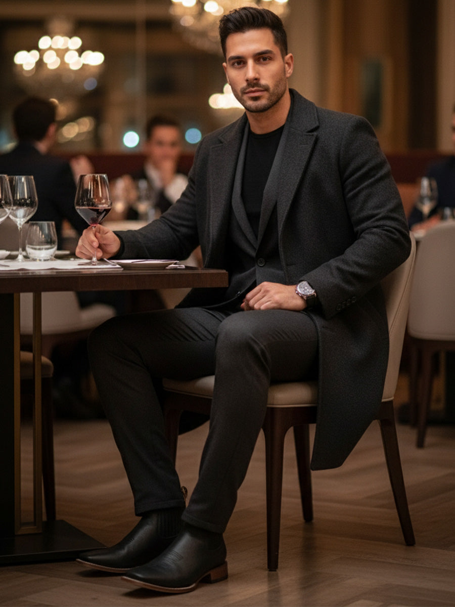 Man in a dark coat sitting at a table in a restaurant holding a glass.