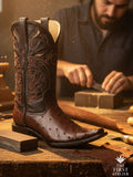 Atelier No. 52 men’s western boot in coffee ostrich leather resting on a wooden workbench, showing rich quill texture and hand-embroidered shaft inside the atelier.
