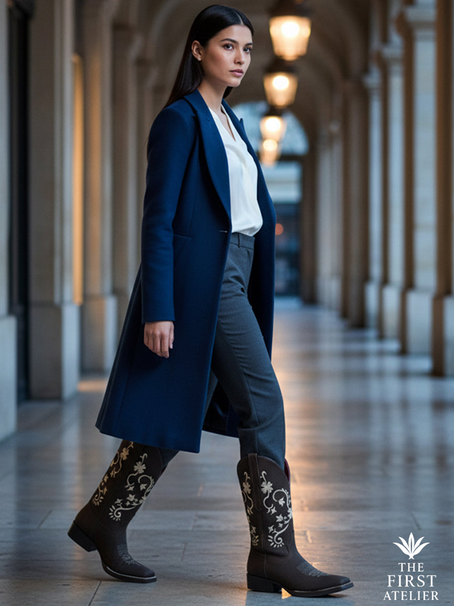 Woman walking under archways at dusk in a navy coat and tailored trousers, wearing La Flor de la Medianoche Boot — handcrafted Mexican leather, Atelier No. 18.