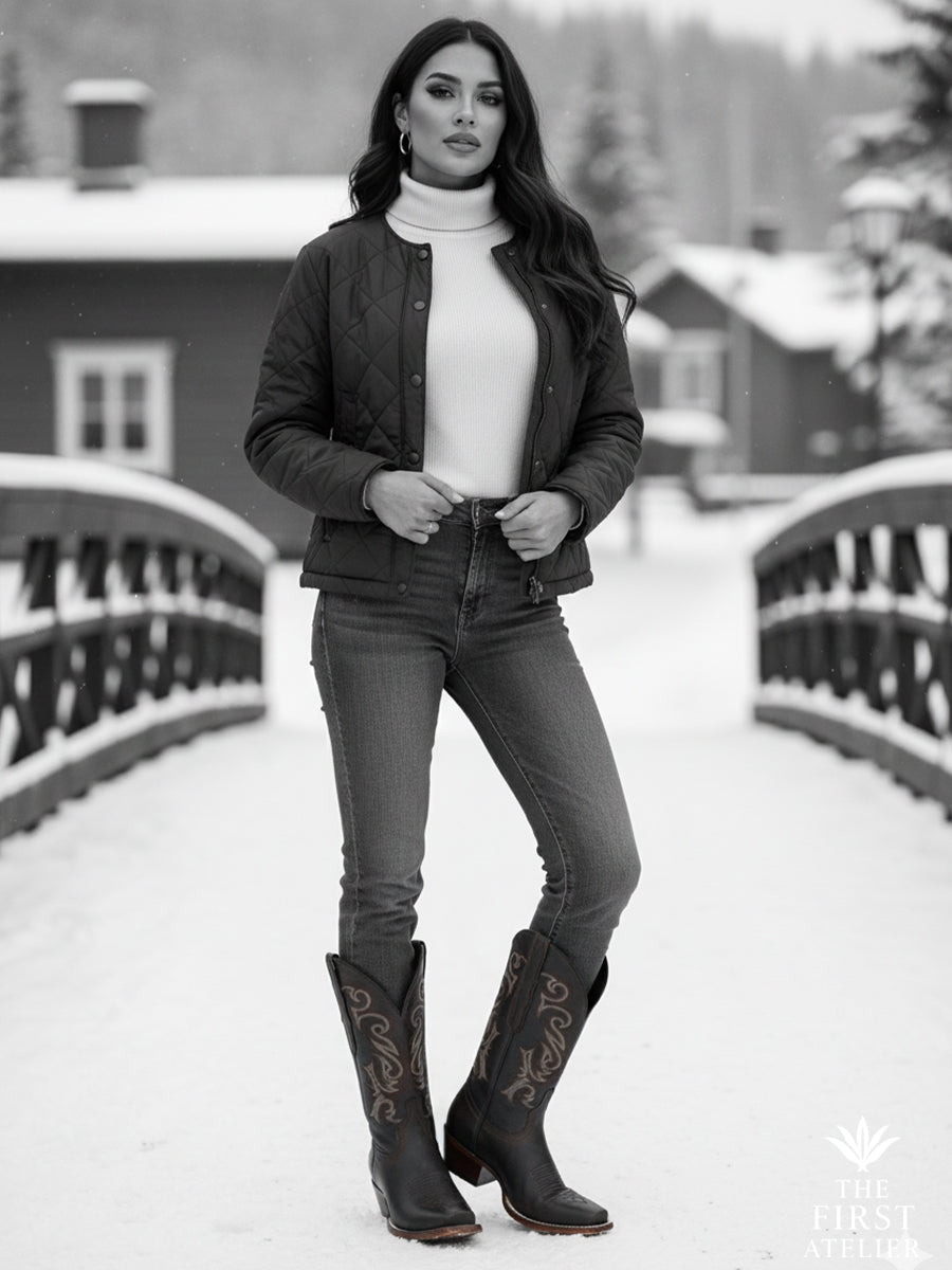 Black-and-white portrait of a woman in the La Ruta del Invierno Boot crossing a snow-covered bridge — quiet confidence framed in winter grace.