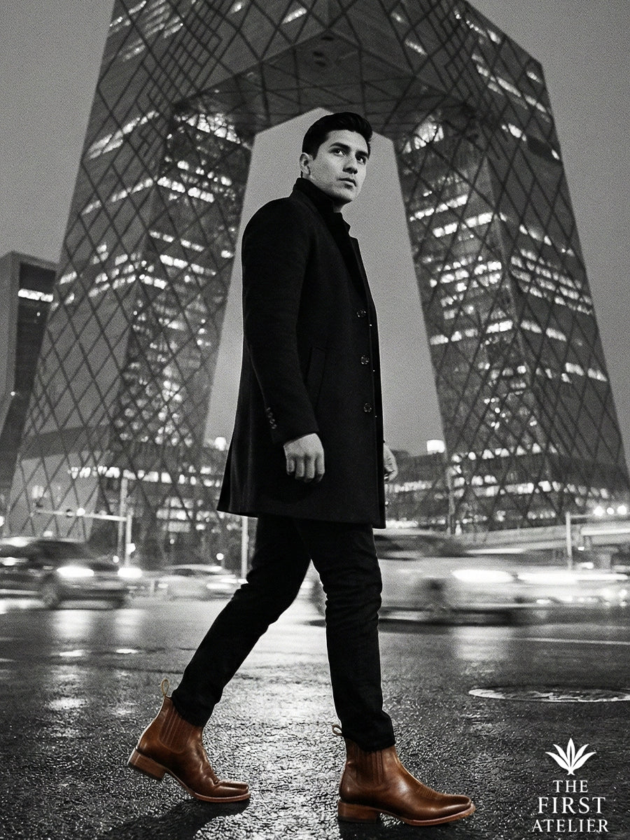 Black and white editorial silhouette of a man in a long coat wearing Atelier No. 68 El Trazo Maestro Boot against a night skyscraper.