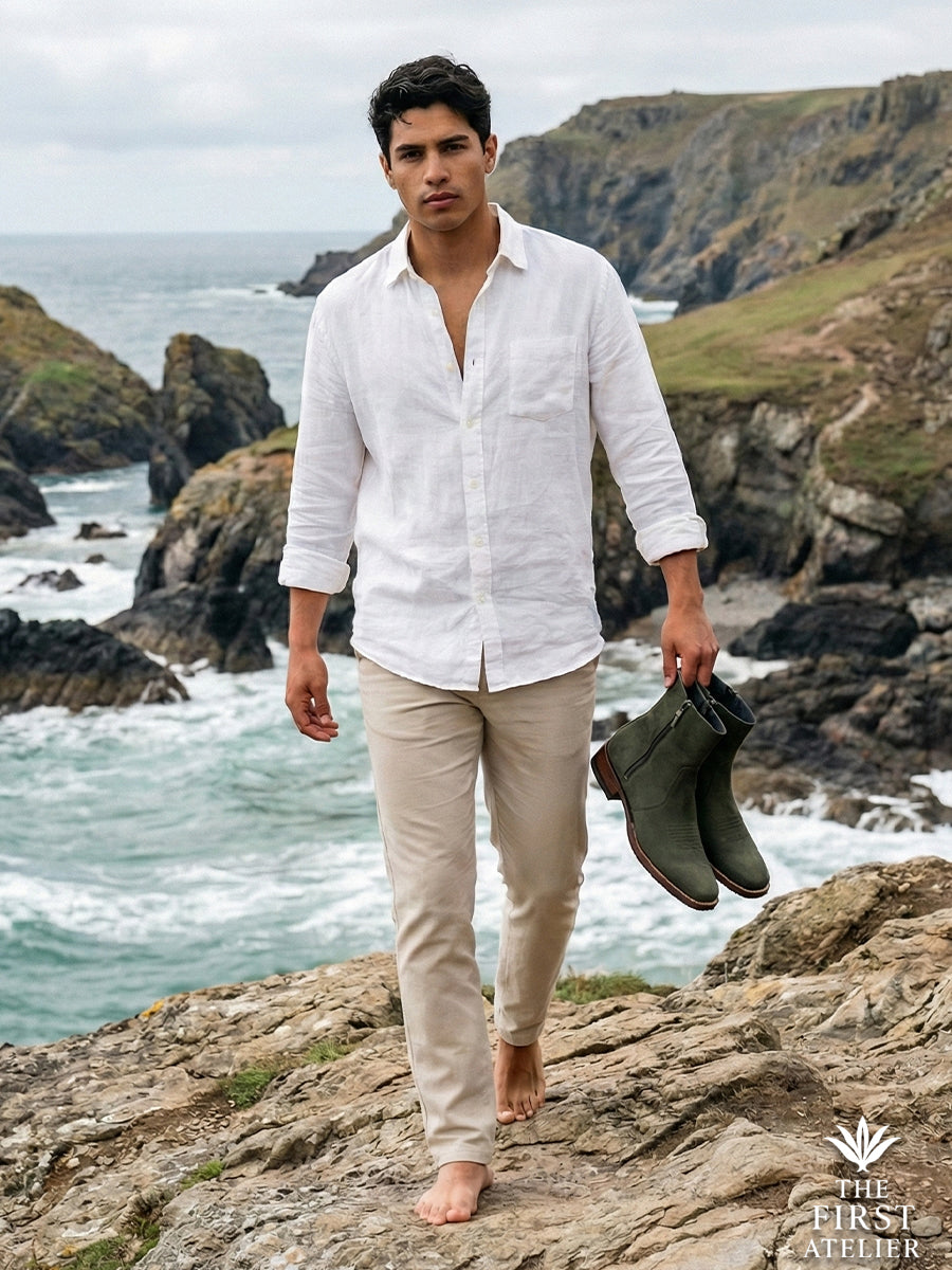 Man walking barefoot on rugged coastal cliffs carrying his Atelier No. 70 El Dominio Boots, showcasing the connection to nature and earth tones.