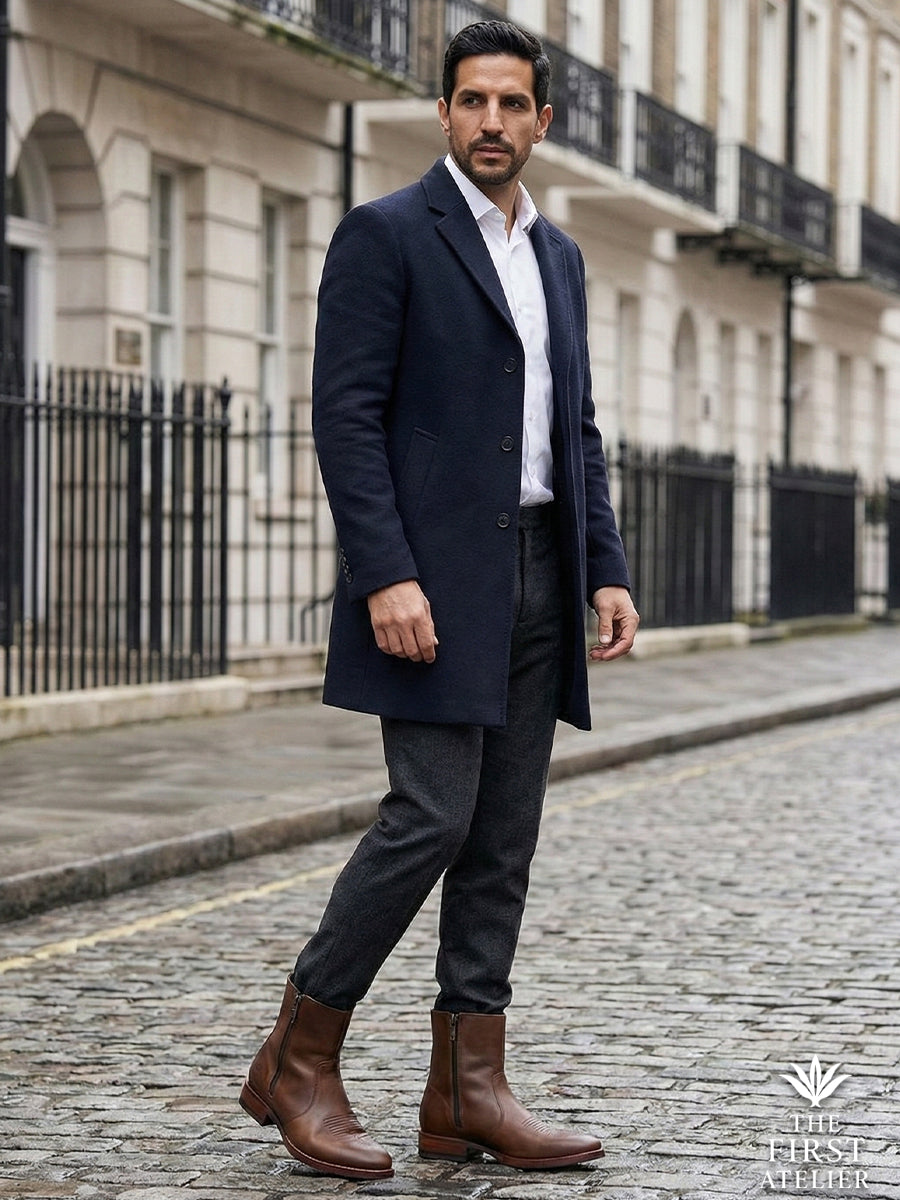 Man in a long navy coat walking on cobblestone streets, wearing the durable and stylish Atelier No. 71 El Destino Boot in brown leather.