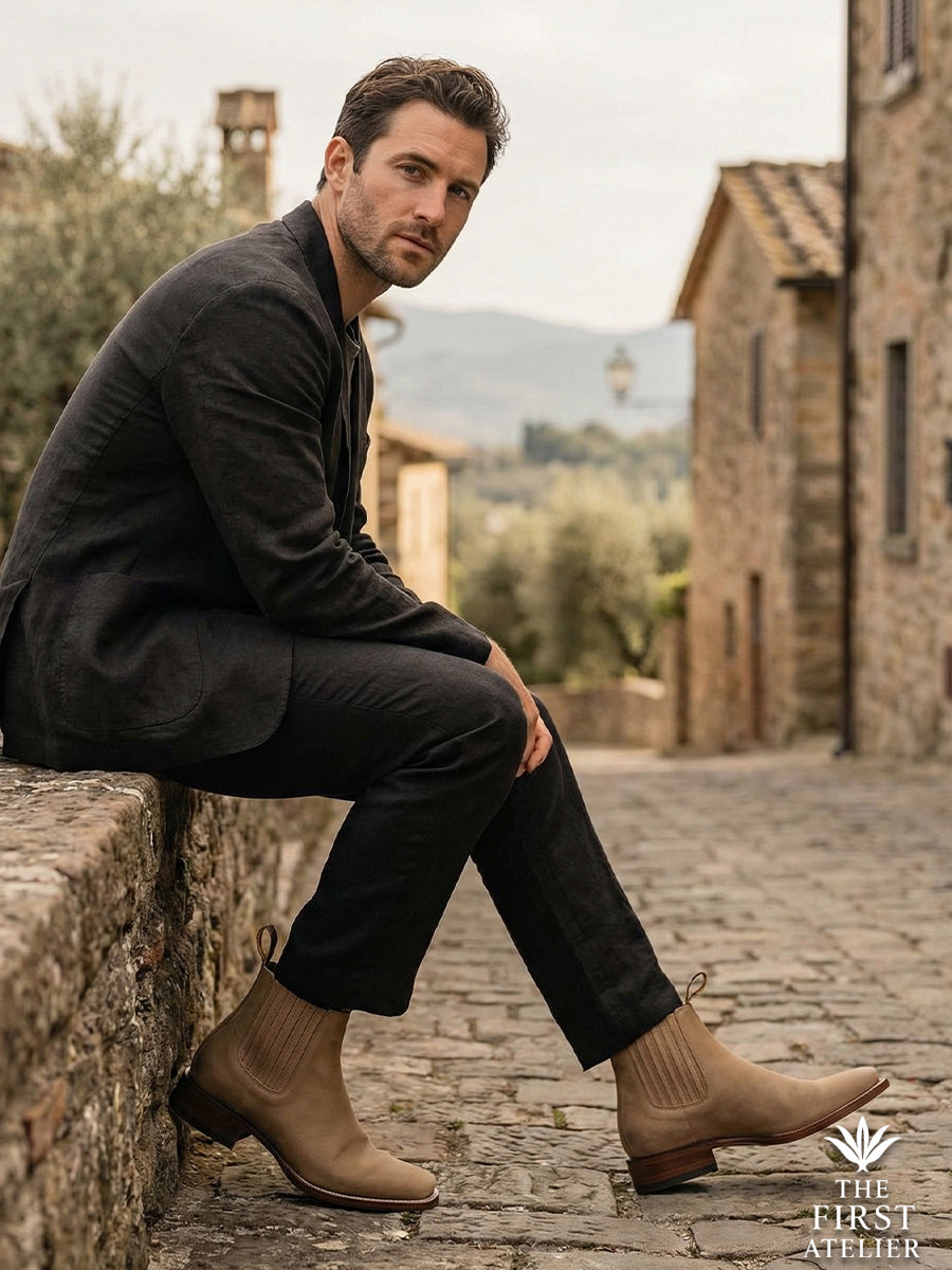 Man resting on a historic stone wall in a European village wearing Atelier No. 73 El Sendero Boot in light tan suede with dark trousers.