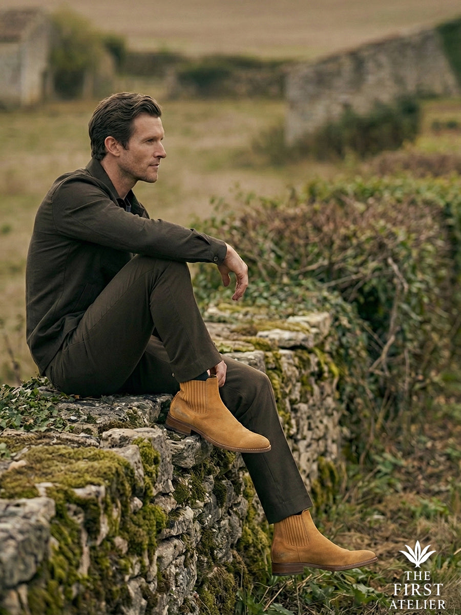 Man sitting on a dry stone wall in the countryside wearing the rugged yet refined Atelier No. 74 Chelsea boot in golden suede.