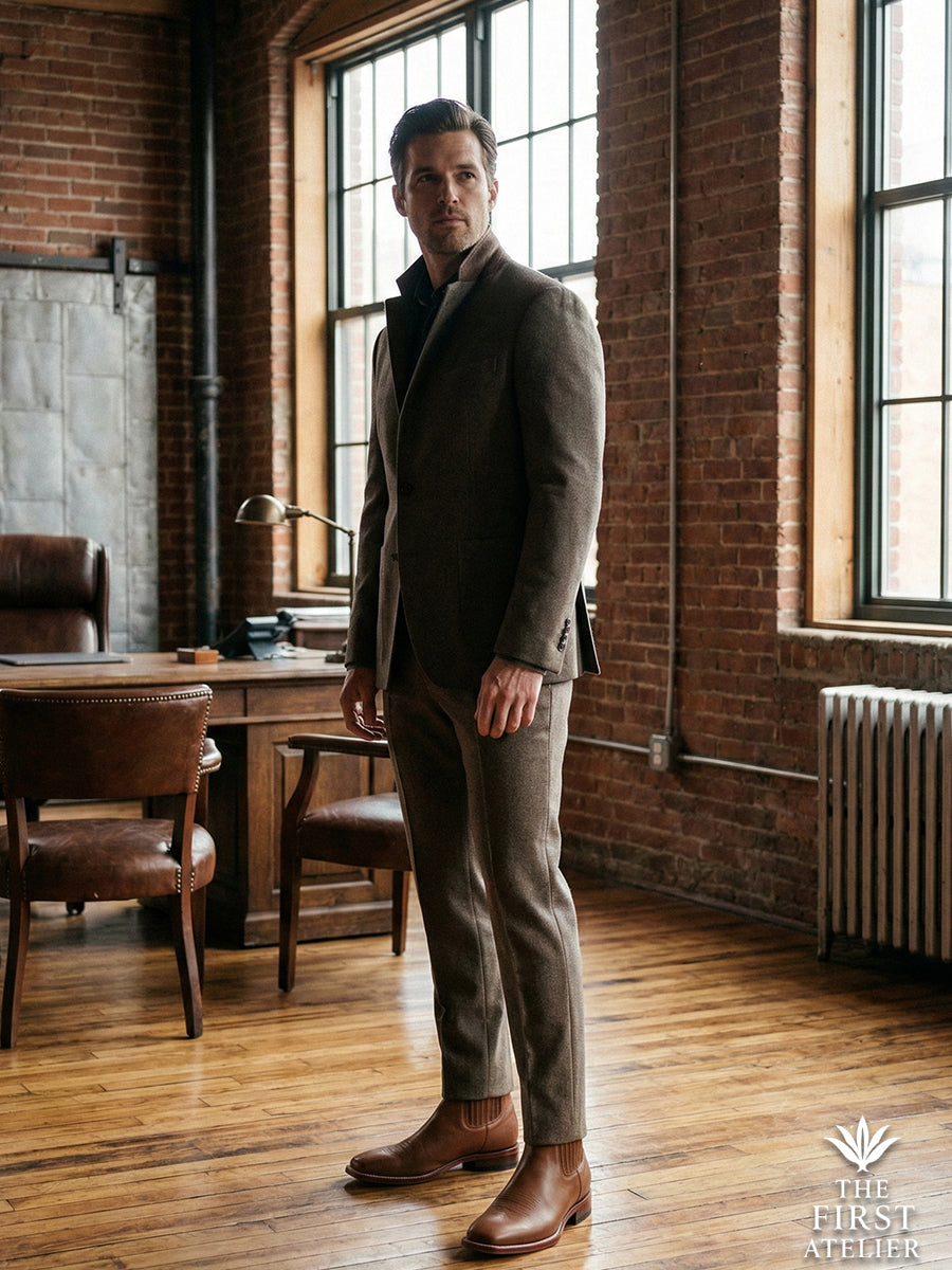 Modern professional style: Man standing in a brick-walled studio wearing grey trousers and the saddle tan Atelier No. 75 Chelsea boot.