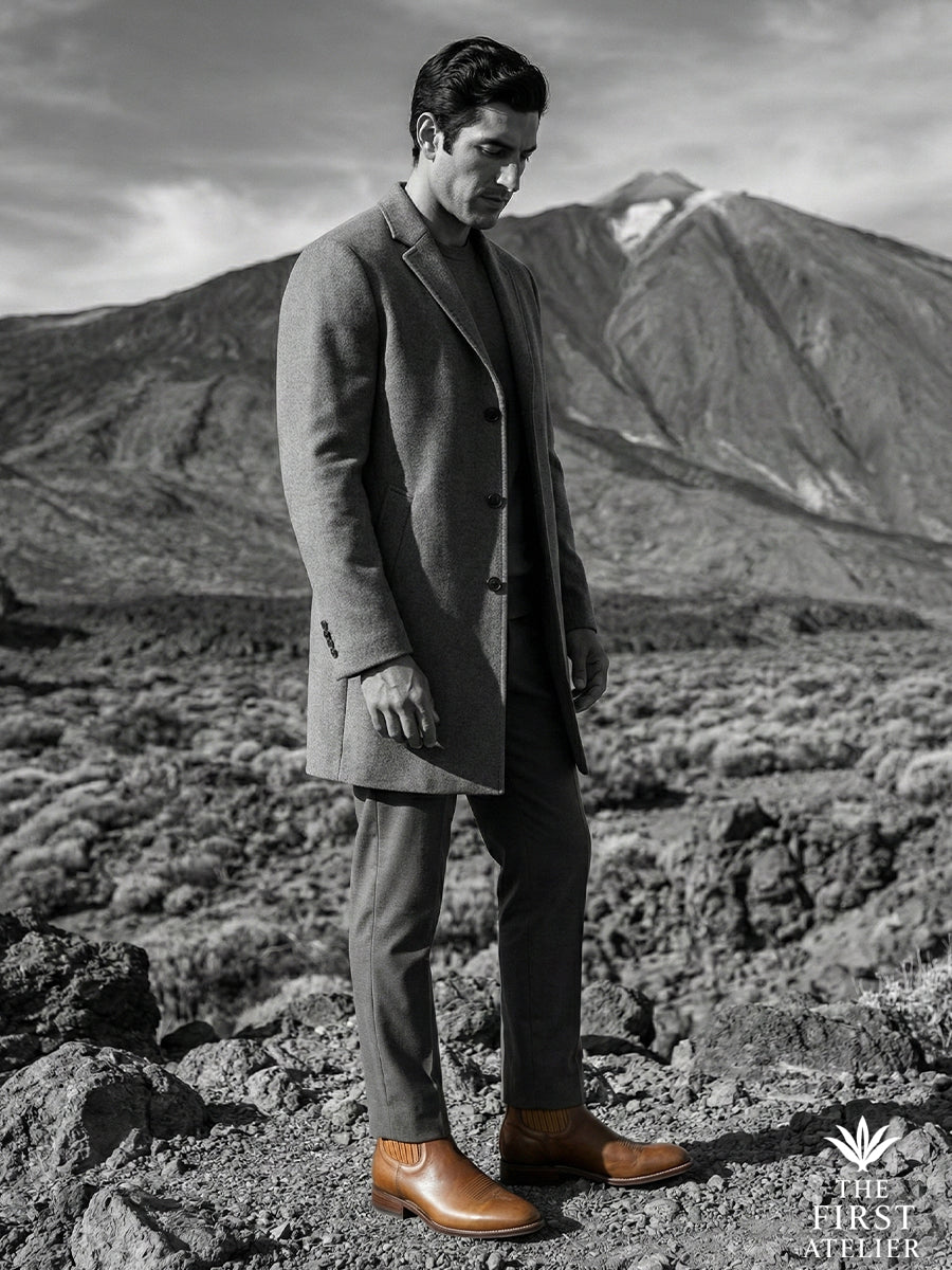 Black and white editorial shot of a gentleman in a long coat standing on rugged terrain, wearing the durable Atelier No. 75 boot.