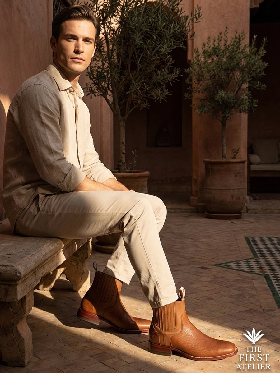 Man resting on a stone bench under an olive tree, embodying relaxed luxury with the textured Atelier No. 77 El Atlas boot.