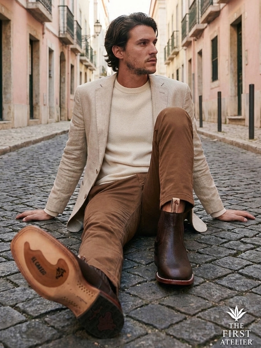 Man sitting on cobblestones in a historic European city wearing beige linen and the dark textured Atelier No. 78 El Alcázar Boot.