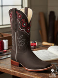 Boot resting on artisan workbench with hand-drawn pattern sheets — chocolate leather with crimson rose embroidery, Atelier No. 25.