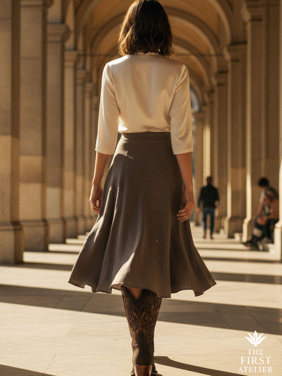 Rear view of the Calle Real Boot under sunlit stone arches — the model in a flowing midi skirt and heels lifted mid-step, evoking quiet power in an Old World corridor, Atelier No. 3.
