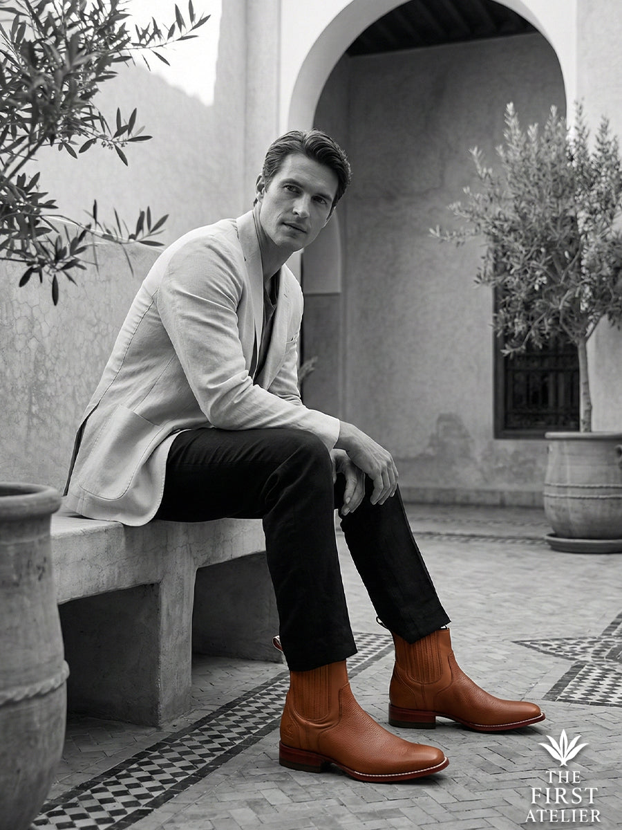 Black and white editorial portrait of a man seated in a historic courtyard, wearing a blazer and the distinctively textured Atelier No. 77 boot.