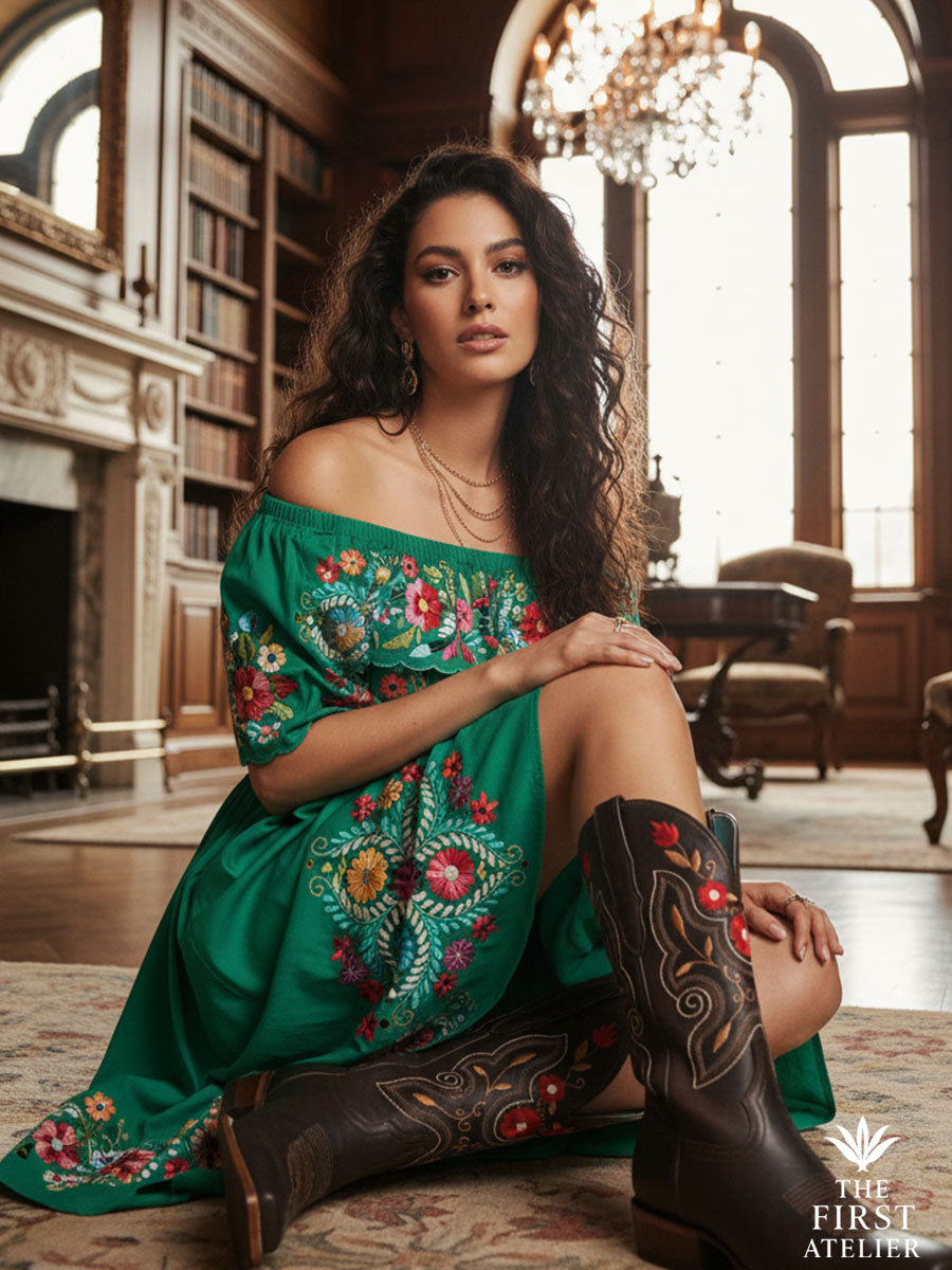 Latina woman seated in a grand interior wearing La Flor de Fuego Boot with an off-shoulder emerald dress, showcasing vibrant red-and-gold embroidery that celebrates Mexican artistry, Atelier No. 02.