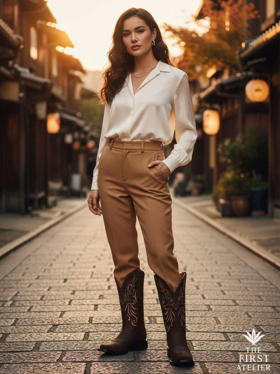 Woman in a white blouse and tailored camel trousers walking a lantern-lit street at dusk in La Rosa del Atardecer Boot — handcrafted Mexican leather, quiet confidence, Atelier No. 14.