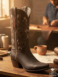 The La Senda Firme Boot in deep brown leather with tonal embroidered western shaft, photographed on the artisan’s worktable surrounded by tools and sketches, Atelier No. 32.
