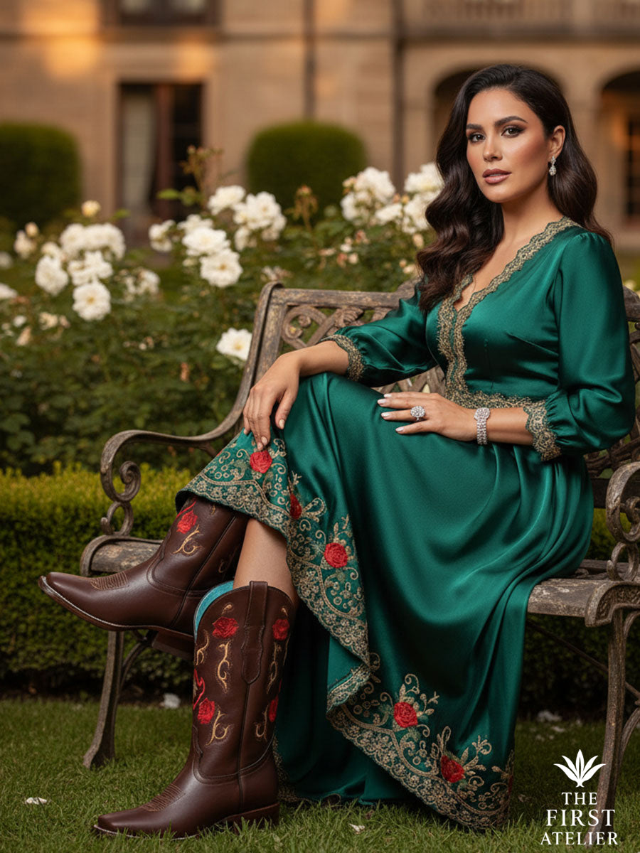 Elegant Latina woman seated in a garden wearing the Rosewood Whisper Boot — handcrafted rosewood leather cowboy boots with crimson and gold floral embroidery — paired with an emerald silk dress, embodying timeless Mexican artistry and poise, Atelier No. 01.v